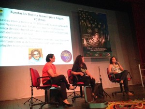 Em um palco três mulheres sentadas em cadeiras vermelhas, a da direita fala ao microfone e as outras a observam. Ao fundo slide da Fundação Dorina projetado e banner do evento.