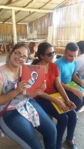 Três adolescentes sentados folheando livros da coleção regionais. EM primeiro plano uma jovem sorrindo exibe o livro Negrinho do Pastoreio.