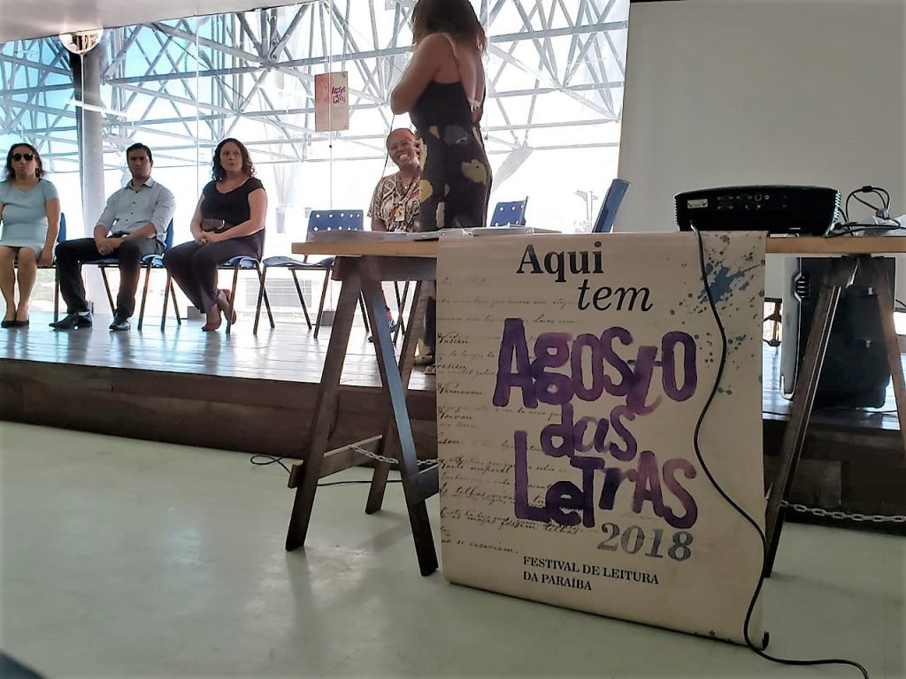 Descrição da imagem: foto de quatro pessoas sentadas, olhando para uma mulher de pé. Ao lado direito há uma mesa com um cartaz escrito "Aqui tem Agosto das Letras 2018 - Festival de Leitura da Paraíba"