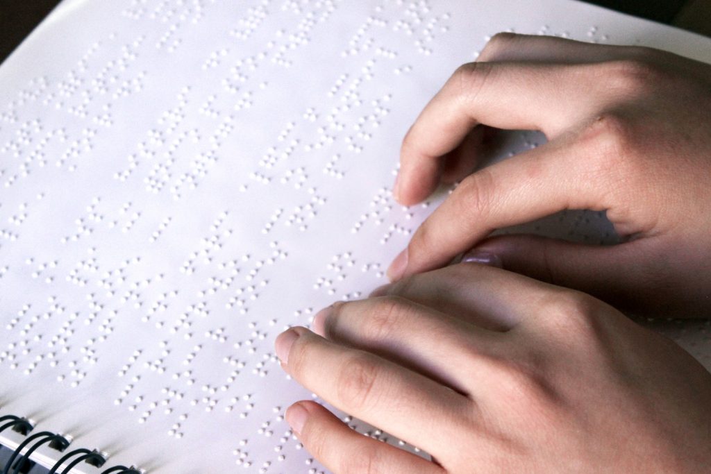 Descrição de imagem: foto de duas mãos sobre uma página em braille.