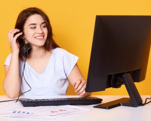 Moça branca sorridente segurando um fone de ouvido com a mão direita à frente de uma computador aberto